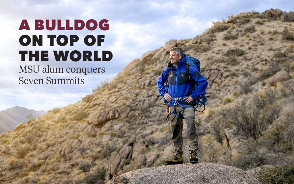 A man in hiking gear stands on a rocky hillside with mountains in the background, under a cloudy sky. Text reads: "A Bulldog on top of the world. MSU alum conquers Seven Summits.