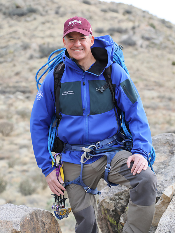 A man in outdoor gear and climbing equipment smiles while posing on rocks in a dry, hilly landscape.