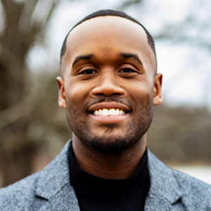 A man with short hair and a trimmed beard smiles outdoors, wearing a grey blazer and black shirt. Trees and an overcast sky are blurred in the background.