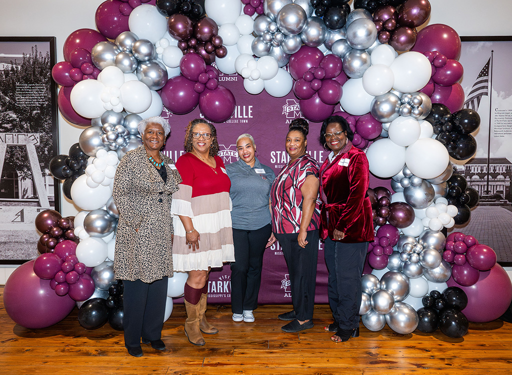 Five women stand in front of a maroon, white, and silver balloon arch and a maroon backdrop with "STARKVILLE" and "ALUMNI" logos.