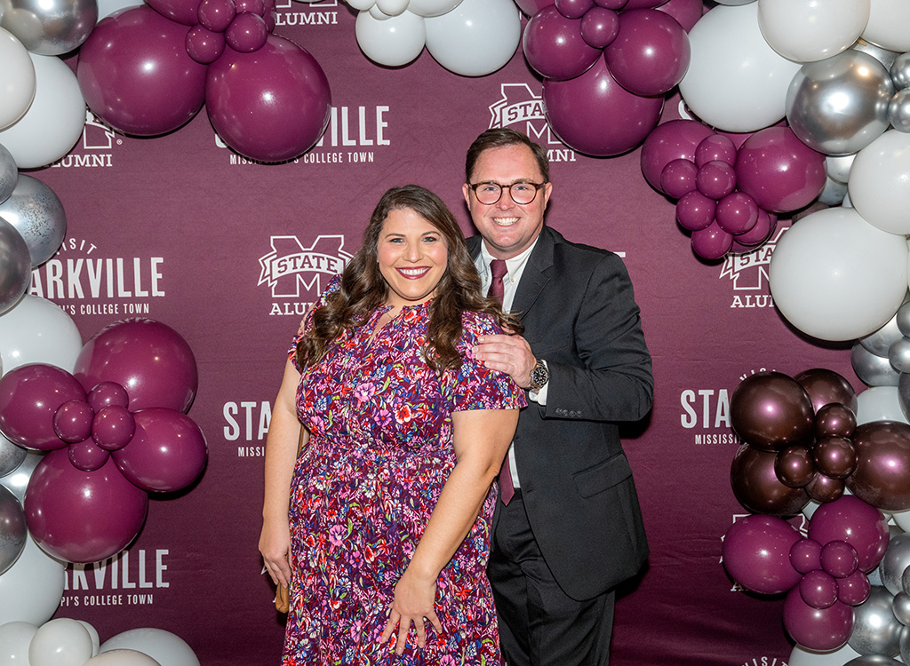 A woman in a colorful dress and a man in a suit pose together in front of a maroon and white balloon arch and a Mississippi State Alumni backdrop.
