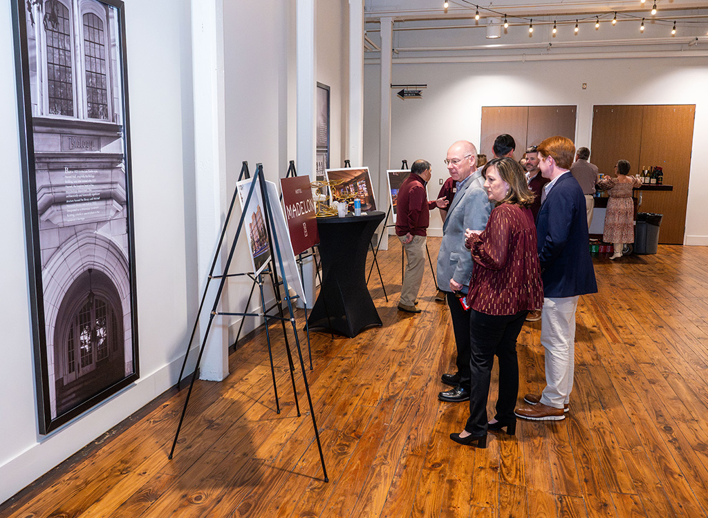 People view framed photographs displayed on easels in a gallery with wooden floors and white walls; others gather near tables in the background.