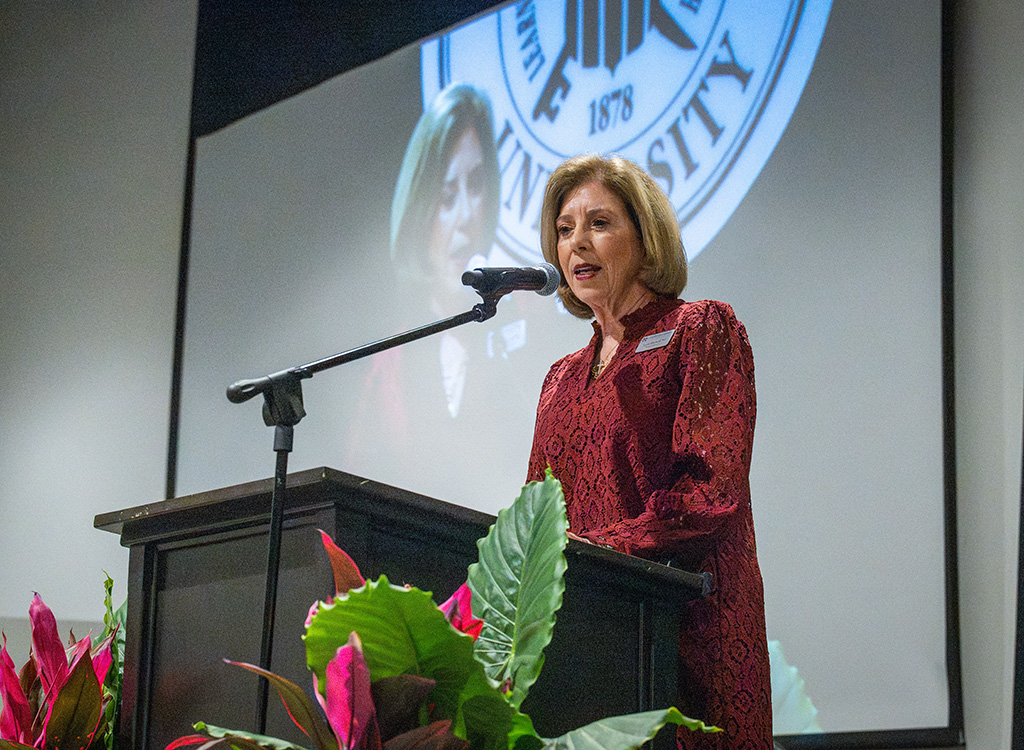 A woman speaks at a podium with a university emblem projected behind her and plants in the foreground.