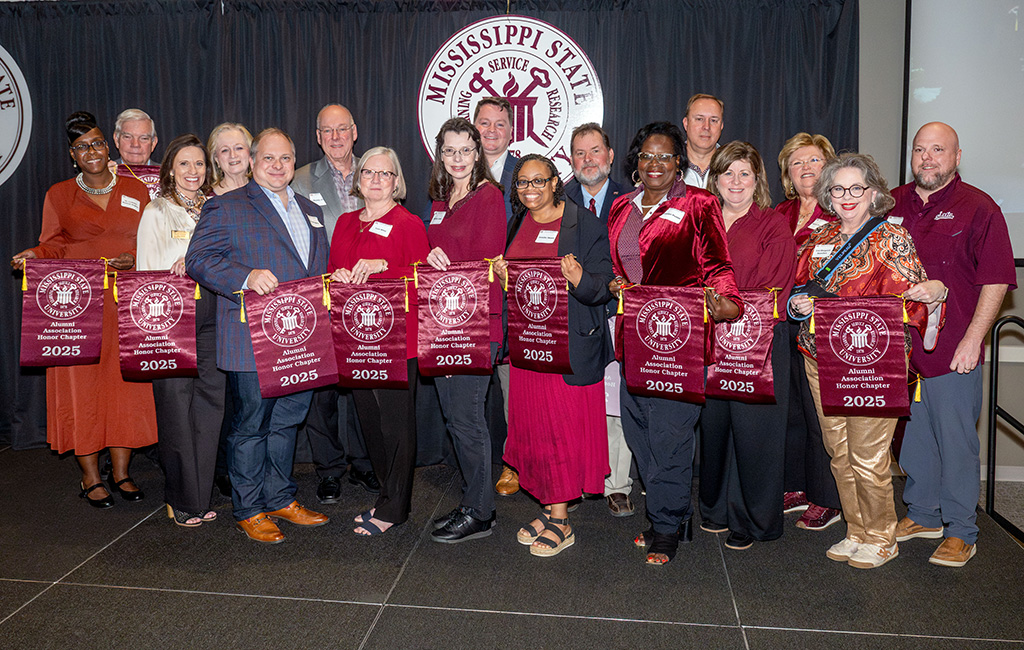 A group of people stands indoors holding maroon banners with “Mississippi State University 2025” written on them, posing for a group photo in front of an MSU backdrop.