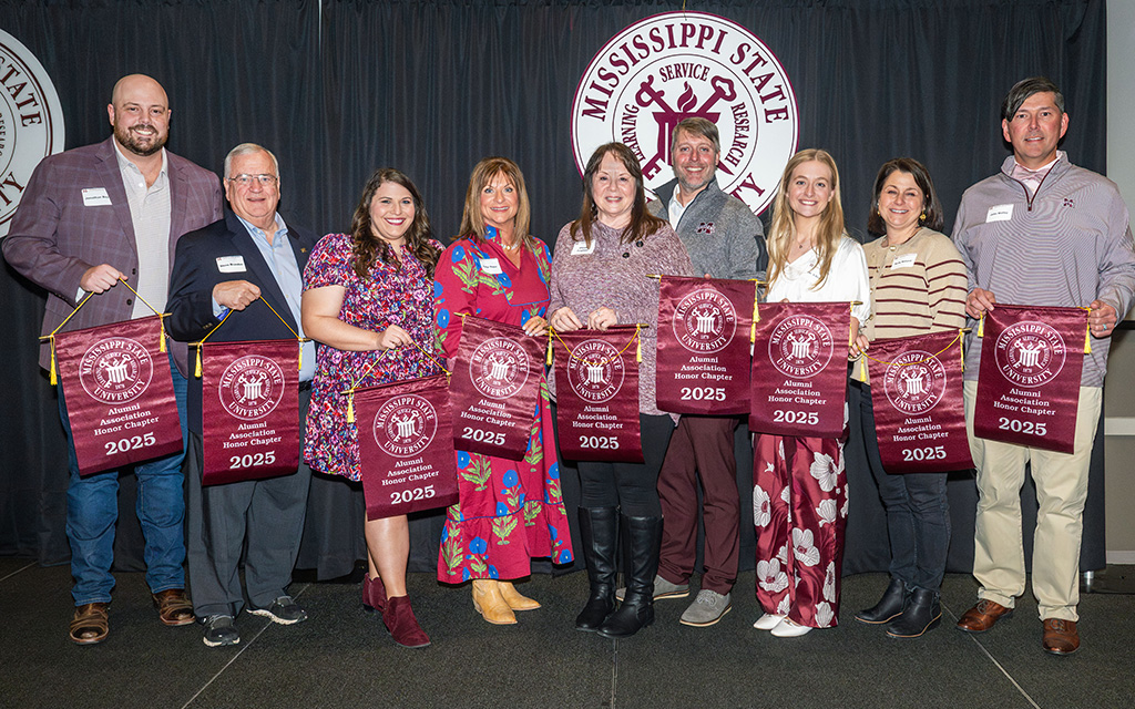 Ten people stand in a row holding maroon banners that read “Mississippi State University Family of the Year 2025,” with a university logo backdrop behind them.