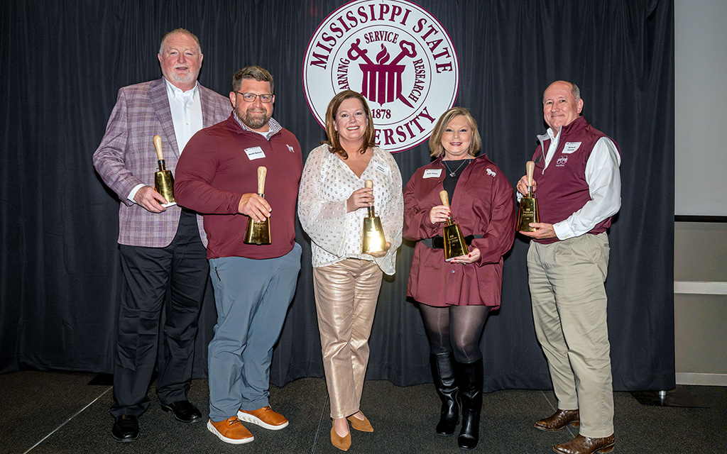 Five people stand in front of a Mississippi State University sign, each holding a large gold bell and smiling at the camera.