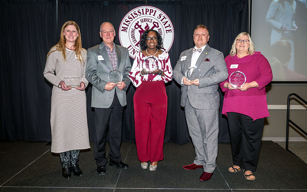 Five people stand on stage holding glass awards in front of a Mississippi State University emblem, posing for a group photo at an event.