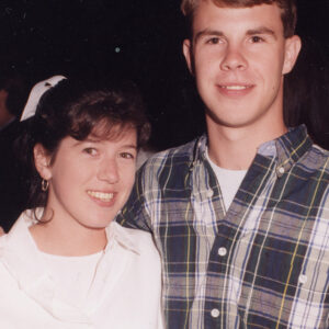A young woman and man stand close together, smiling at the camera. The woman wears a white shirt, and the man wears a plaid button-up shirt. The background is dark and out of focus.