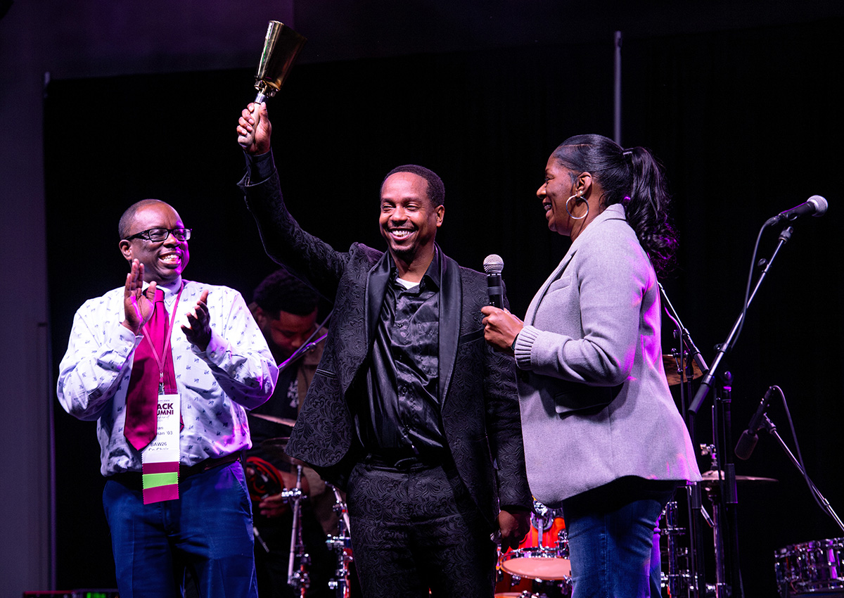 Three people on stage; the person in the center raises a bell, while the others clap and smile. Microphones and musical instruments are visible in the background.