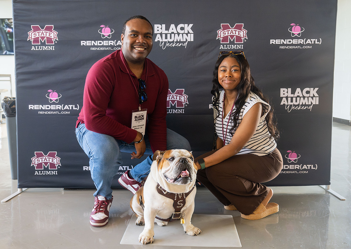 Two people pose with a bulldog in front of a backdrop that reads "Mississippi State Alumni" and "Black Alumni Weekend.
