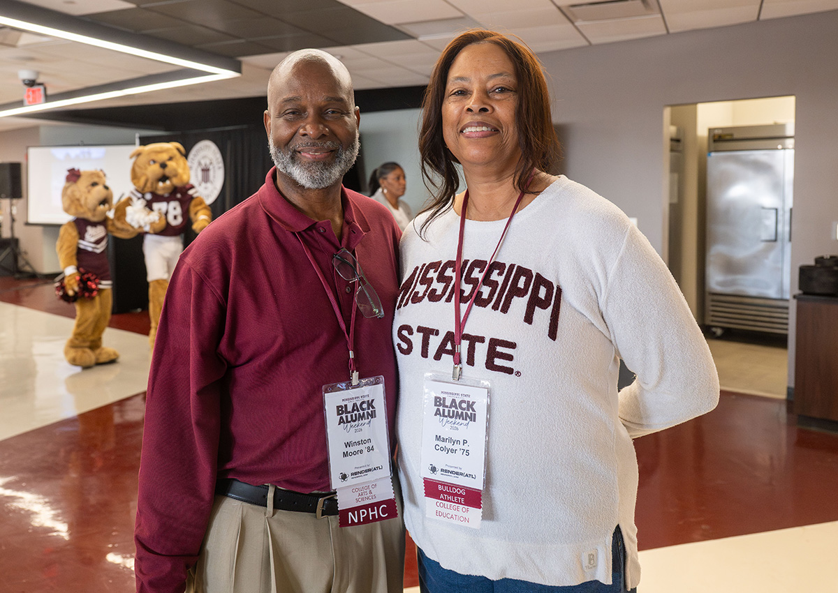 A man and woman wearing Mississippi State attire stand indoors at a Black Alumni event; two mascots are visible in the background.