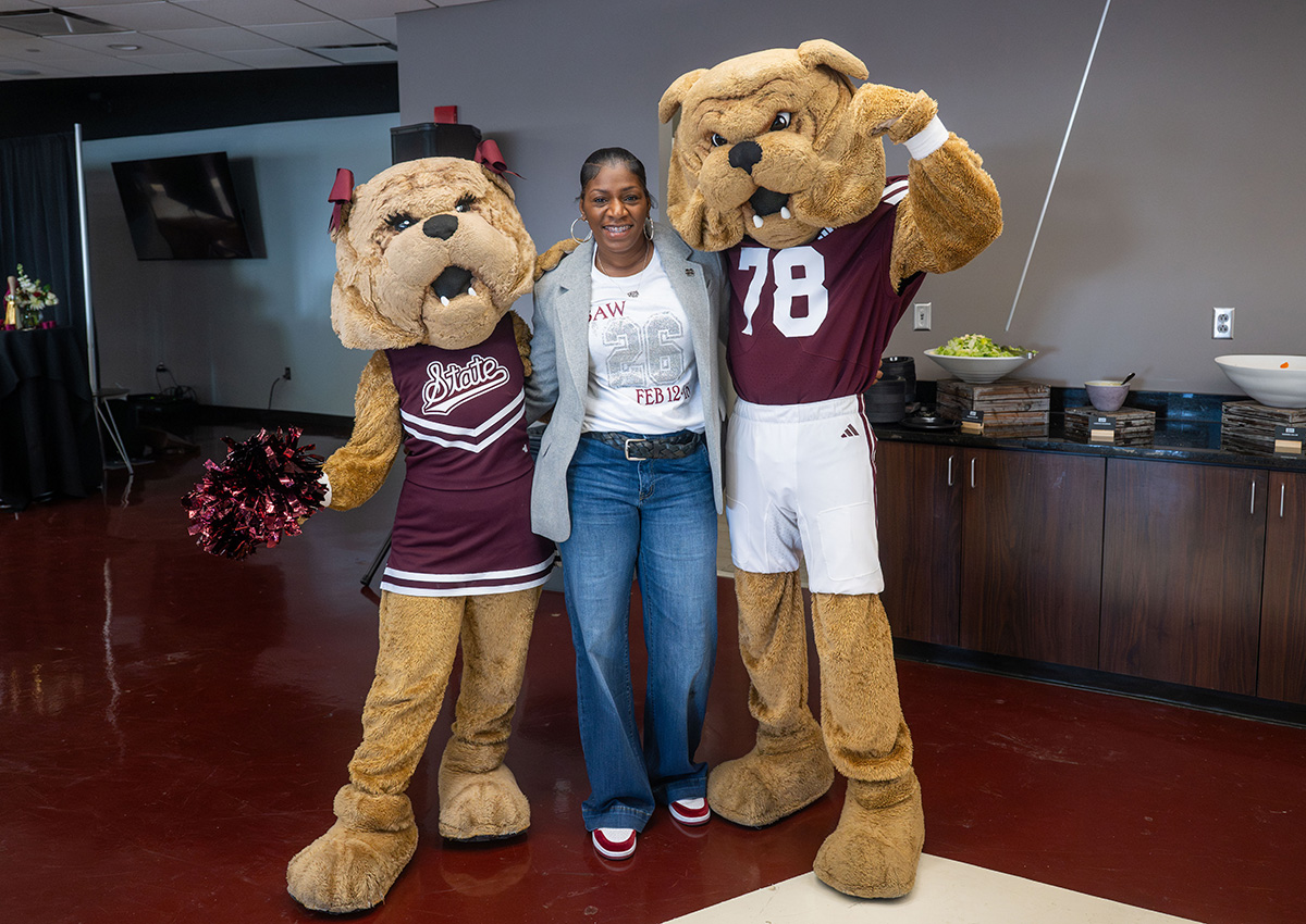 A woman stands between two bulldog mascots, one in a cheerleader outfit and the other in a sports uniform, inside a room with a counter and snacks in the background.
