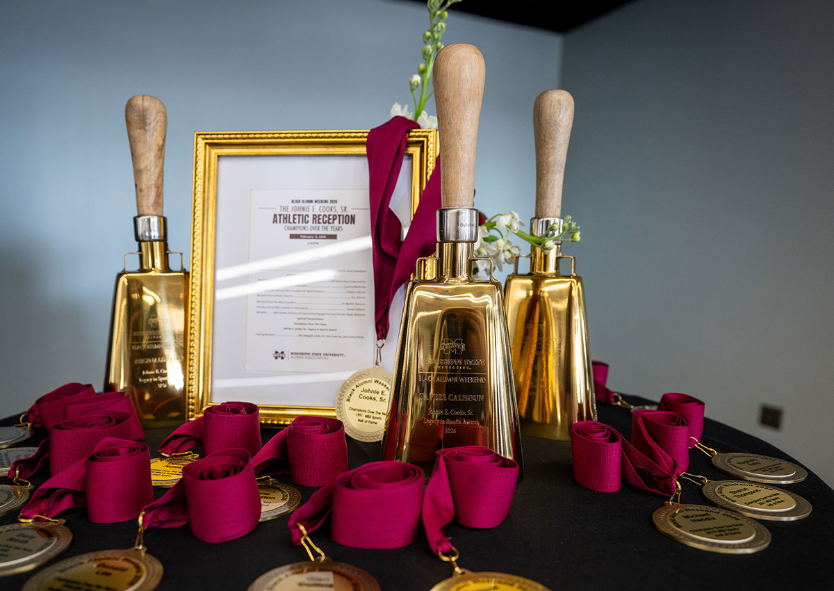 Three gold cowbells with wooden handles and multiple gold medals with maroon ribbons are arranged on a table next to a framed certificate or document.