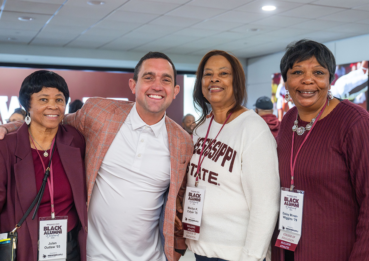 Four people, three women and one man, stand together smiling at a Black alumni event. Two women wear name tags and Mississippi State shirts. Indoor setting with others in the background.