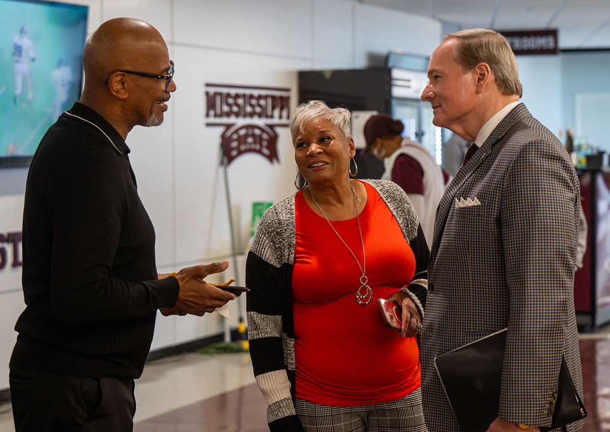 Three adults stand indoors having a conversation; one man holds a laptop, while the other man and a woman listen. Mississippi State signs are visible in the background.