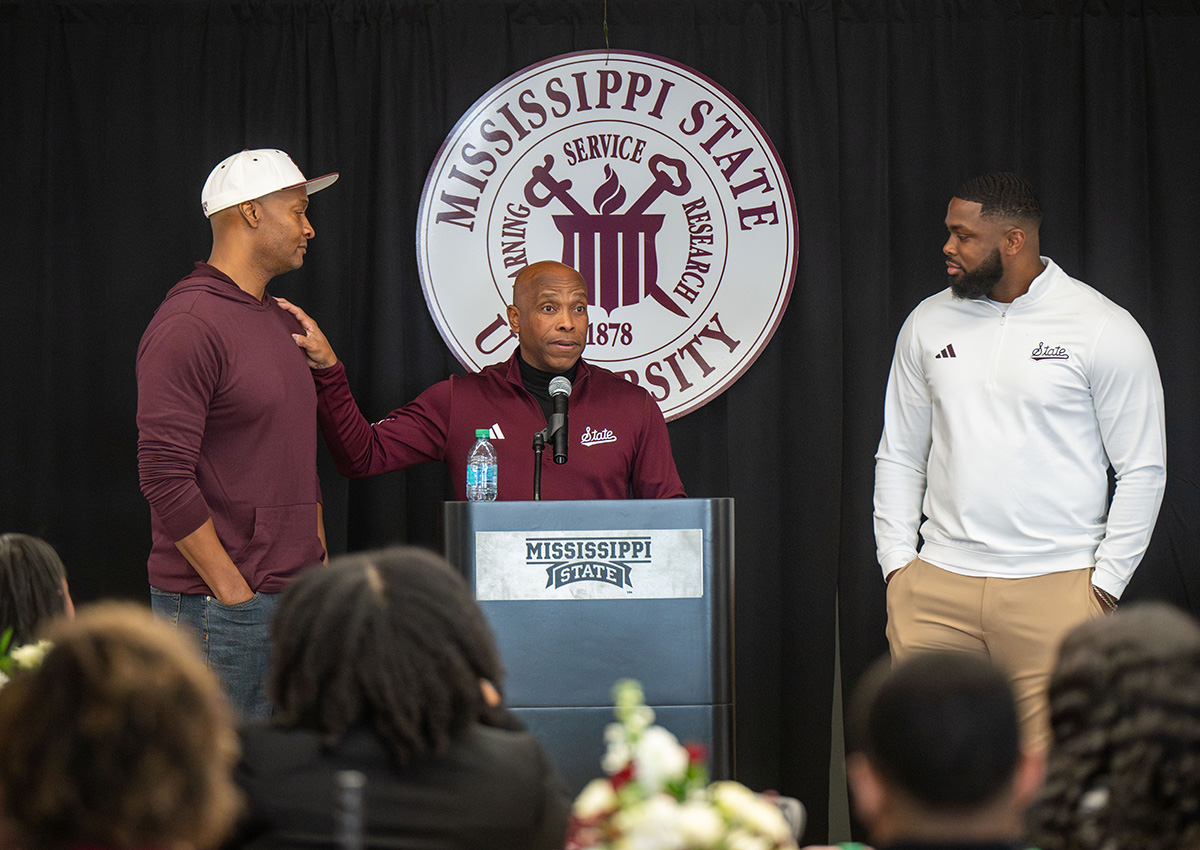 Three men stand on a stage at Mississippi State University, with one speaking at a podium and the university seal visible in the background.