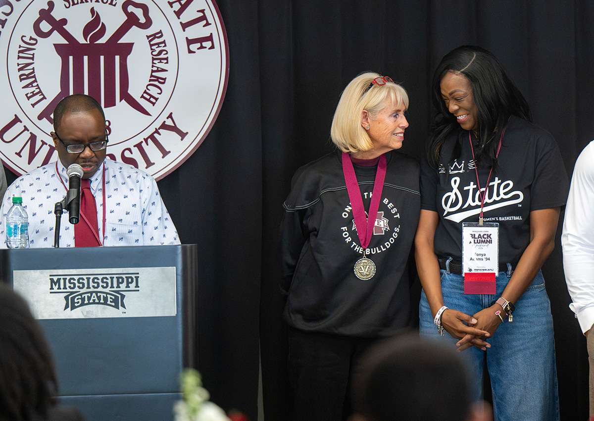 Three people stand on a stage at a Mississippi State University event; two women wearing medals smile at each other while a man speaks at a podium.