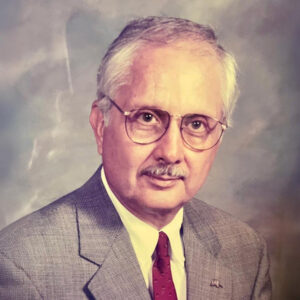 An older man with gray hair and glasses wearing a gray suit, white shirt, and red tie, posing for a formal portrait against a neutral background.
