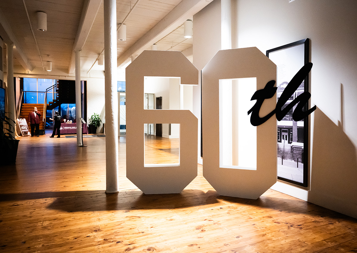 Large cutout reading "60th" displayed in a spacious, modern hallway with wood floors and white walls; framed photos hang in the background.