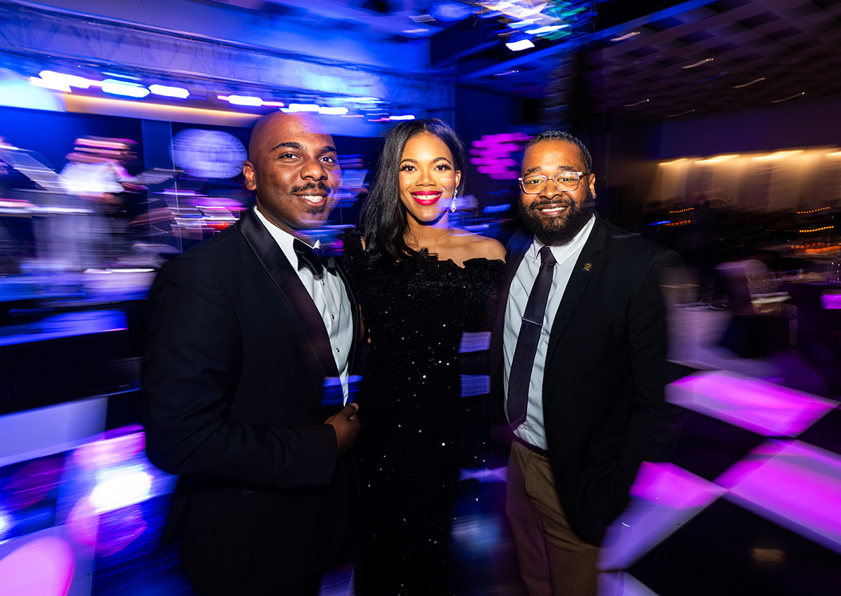 Three people dressed in formal attire stand together and smile for a photo at an indoor event with colorful stage lights in the background.
