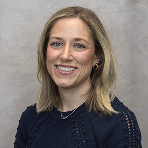 A woman with shoulder-length blonde hair, wearing a navy blue sweater and gold hoop earrings, smiles at the camera against a light gray background.