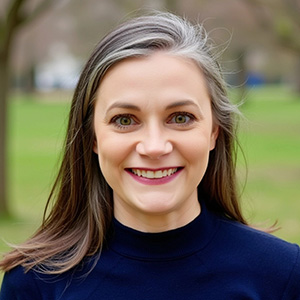A woman with long brown hair and gray streaks, wearing a navy blue top, smiles outdoors with trees and grass in the background.