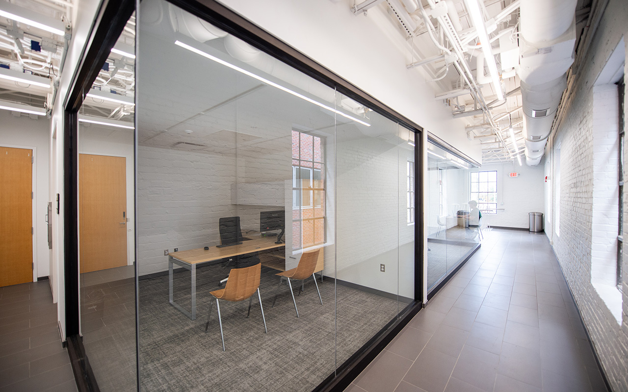 A modern office hallway with glass-walled meeting rooms featuring wooden chairs, computer monitors, tiled floors, and exposed ceiling pipes.