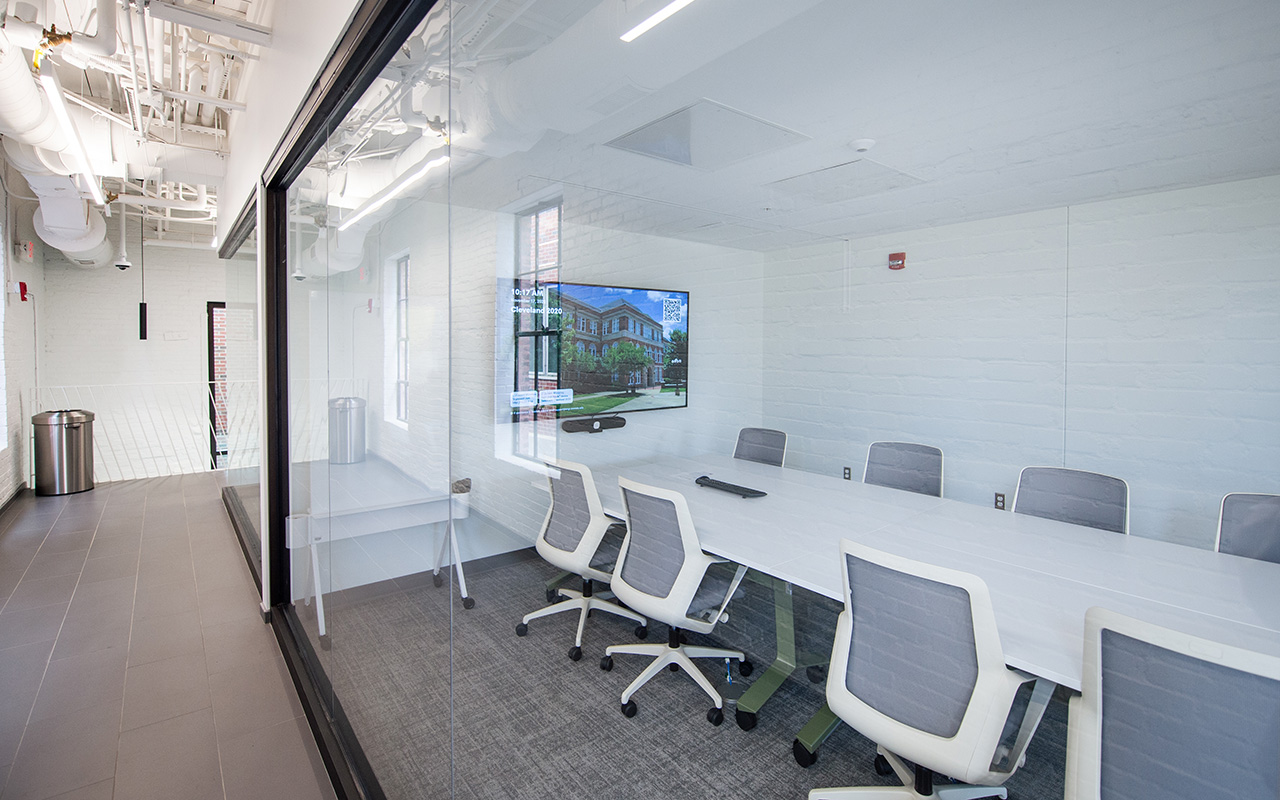 A modern conference room with six white chairs around a white table, a wall-mounted screen, and large glass windows looking into a hallway.