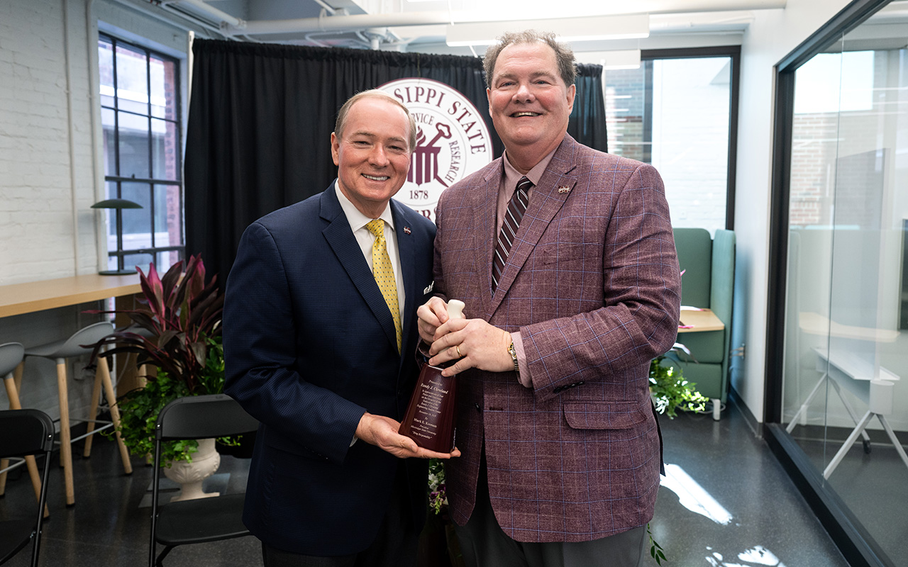 Two men in suits stand indoors, smiling and holding an award together in front of a Mississippi State University backdrop.