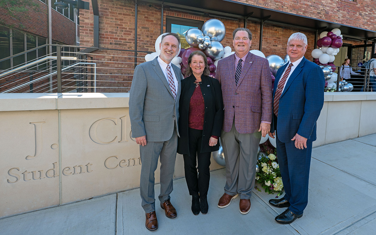 Four adults in business attire stand outside near a brick building and decorative balloons, in front of a sign reading "J. C. Student Center.