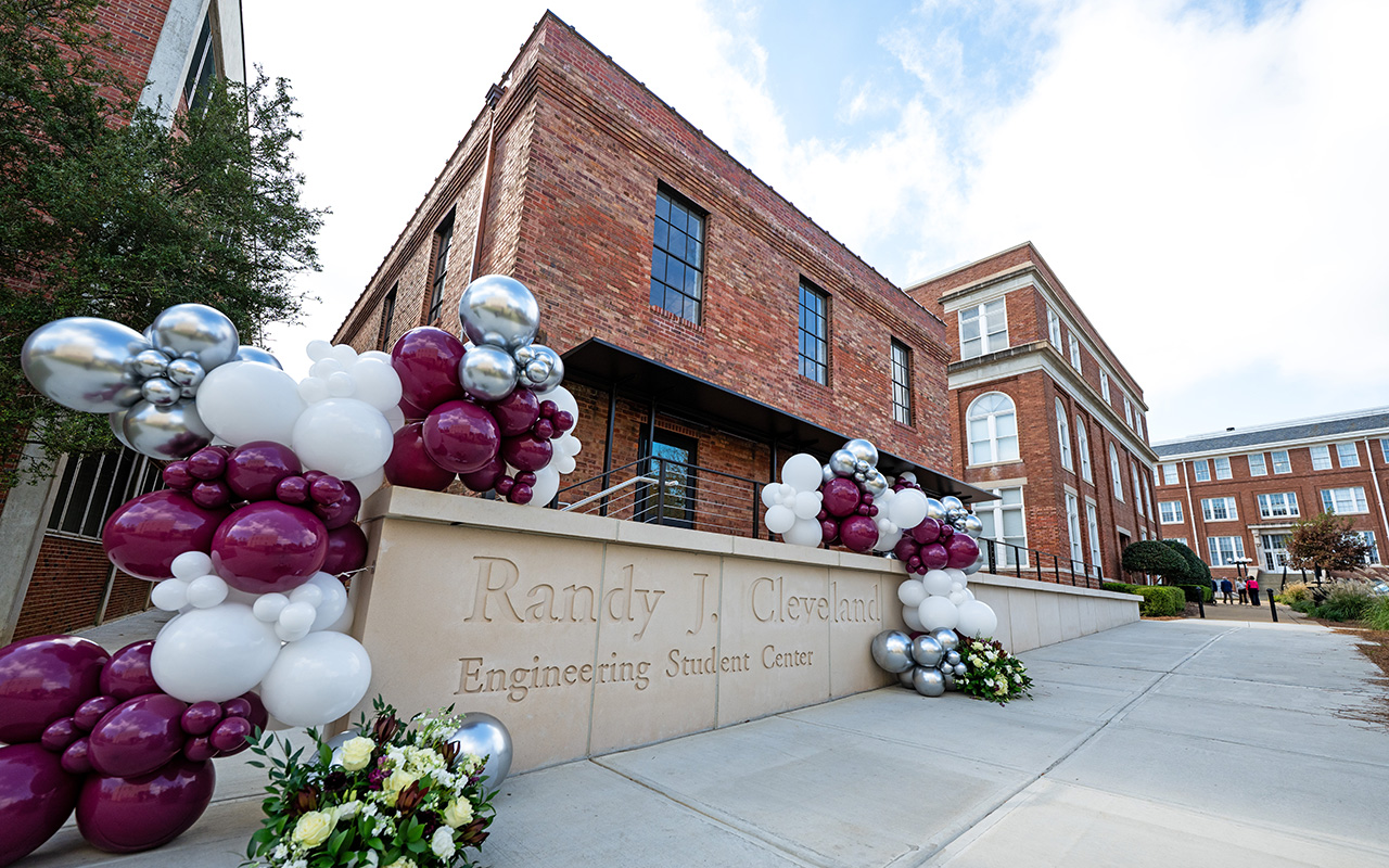 A brick building labeled "Randy J. Cleveland Engineering Student Center" is decorated with clusters of white, maroon, and silver balloons and floral arrangements.