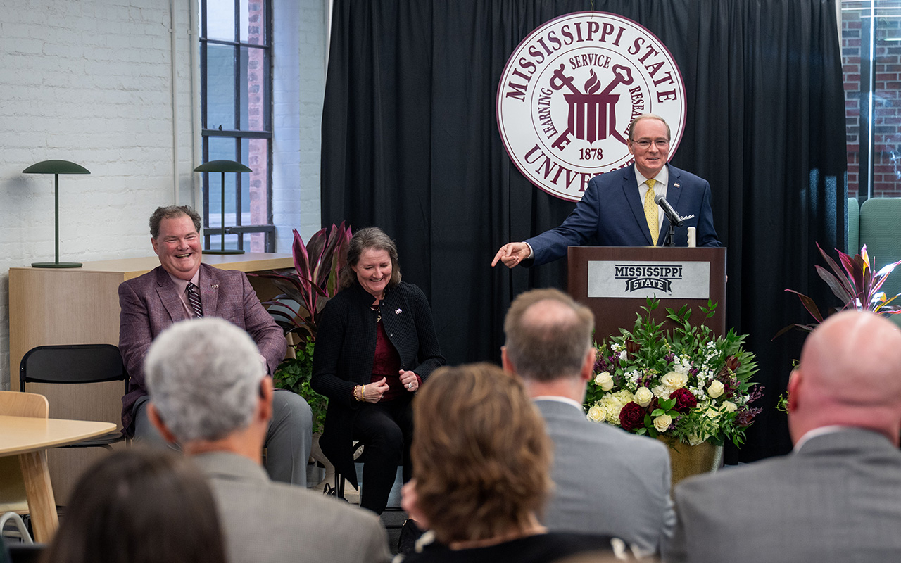 A man speaks at a podium with the Mississippi State University seal behind him while two seated individuals and an audience listen.