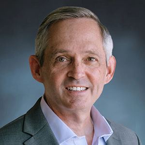 A middle-aged man with short gray hair wearing a gray suit jacket and light blue shirt, smiling in front of a dark, blurred background.