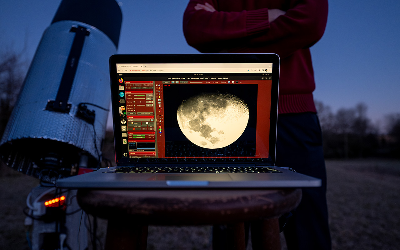 A laptop displaying a close-up image of the moon sits on a stool outdoors near a telescope, with a person standing nearby at dusk.