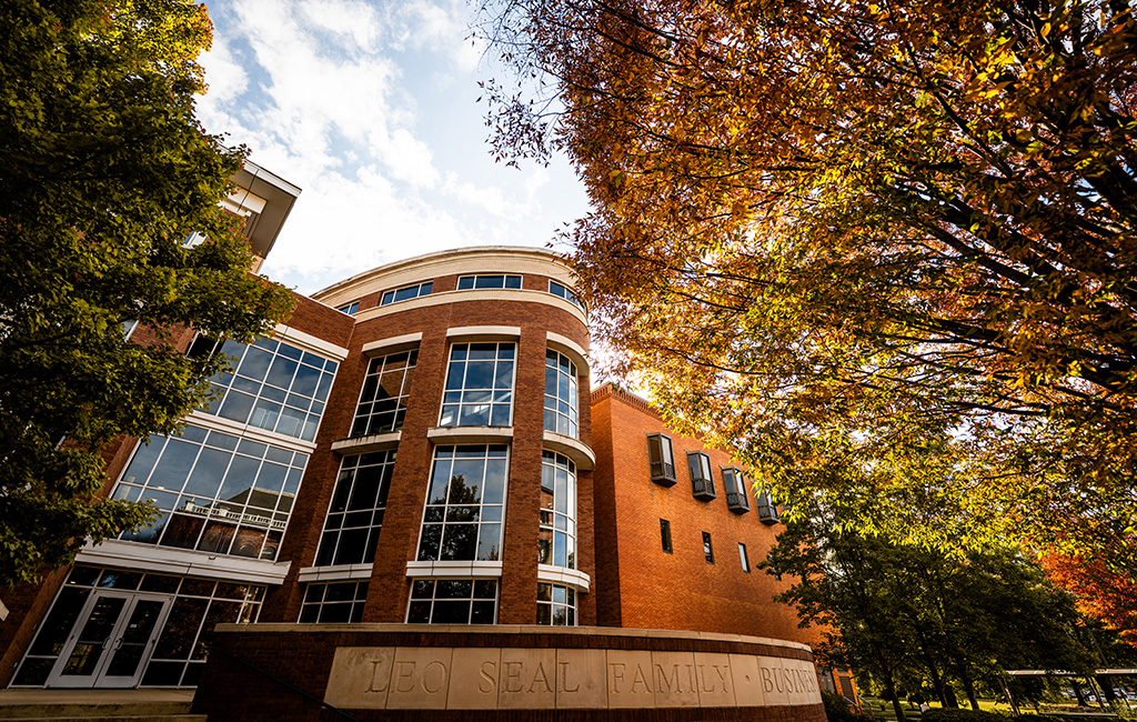 Red-brick academic building with large windows, surrounded by trees with autumn foliage under a partly cloudy sky.