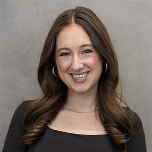 A woman with long brown hair, wearing a black top, gold hoop earrings, and a gold necklace, smiles in front of a gray background.