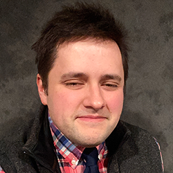 A man with short dark hair wearing a plaid shirt, dark vest, and tie poses in front of a gray textured background.