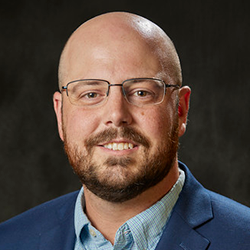 A man with a shaved head, beard, and glasses wearing a blue suit jacket and light blue shirt, smiling in front of a dark background.