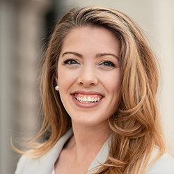 A woman with long, light brown hair smiles at the camera, wearing a light-colored blazer and pearl earrings. The background is blurred.