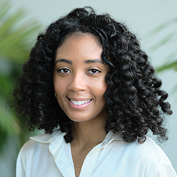 A woman with curly black hair, wearing a white blouse, smiles at the camera. A green plant is blurred in the background.