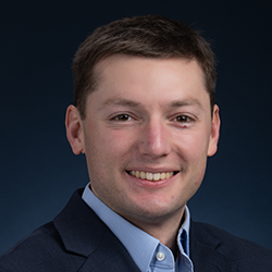 A man with short brown hair, wearing a dark suit jacket and light blue shirt, smiling in front of a dark blue background.