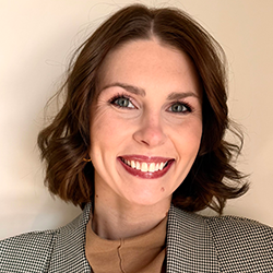 A woman with short brown hair and blue eyes smiles at the camera. She is wearing a checked blazer over a tan top against a light background.