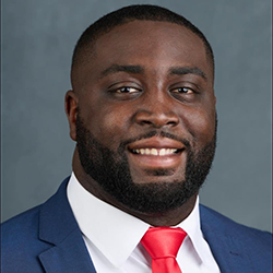 A man in a blue suit, white shirt, and red tie is smiling at the camera against a plain gray background.