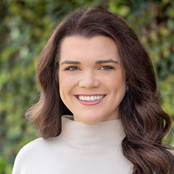 A woman with long brown hair, wearing a light-colored turtleneck, smiles in front of a blurred green leafy background.