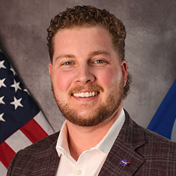 A man with short curly hair and a beard, wearing a plaid blazer and white shirt, stands in front of an American flag and another blue flag.