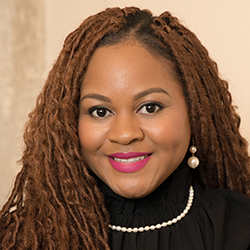 Woman with long brown locs, wearing a black top, pearl necklace, pearl earrings, and bright pink lipstick, smiling at the camera with a neutral background.