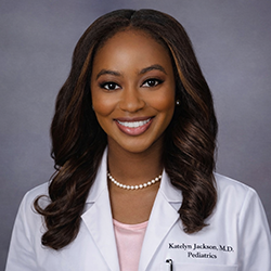 A woman wearing a white lab coat and pearl necklace smiles at the camera against a plain gray background.
