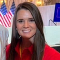 A woman with long brown hair wearing a red shirt smiles indoors. Two American flags and a large screen are visible in the background.