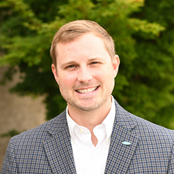 A man in a checked blazer and white dress shirt smiles in front of green foliage.