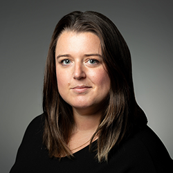 Woman with straight brown hair and a neutral expression wearing a black top, posing against a plain gray background.